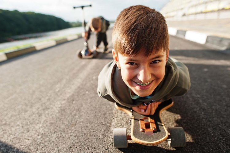 A young boy riding a skateboard on his belly looking up with a big smile with metal braces on his teeth