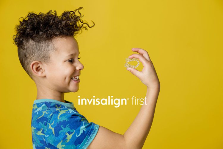 A young boy with a blue, dinosaur t-shirt holding up his Invisalign First aligner while smiling
